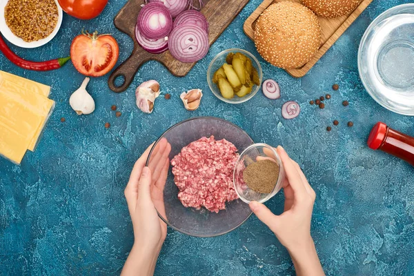Top view of woman adding black pepper to minced meat near burger ingredients on blue textured surface — Stock Photo