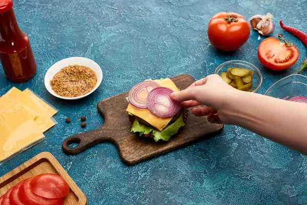 Vista recortada de la mujer poniendo cebolla en hamburguesa fresca en la superficie de textura azul - foto de stock