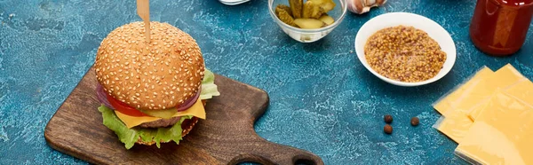 Fresh cooked burger on wooden cutting board near ingredients on blue textured surface, panoramic shot — Stock Photo