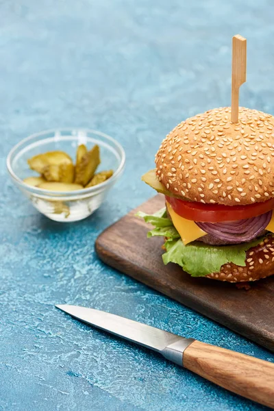 Selective focus of delicious cheeseburger on wooden board near pickles and knife on blue textured surface — Stock Photo