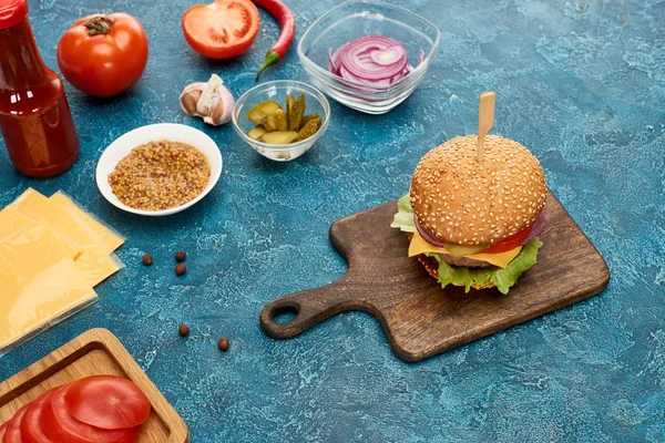 Fresh cooked burger on wooden cutting board near ingredients on blue textured surface — Stock Photo