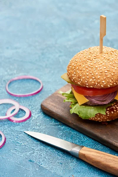 Delicious cheeseburger on wooden board near knife on blue textured surface — Stock Photo