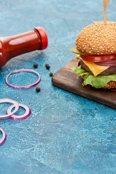 Deliciosa hamburguesa con queso sobre tabla de madera cerca de cebolla y ketchup sobre superficie de textura azul - foto de stock