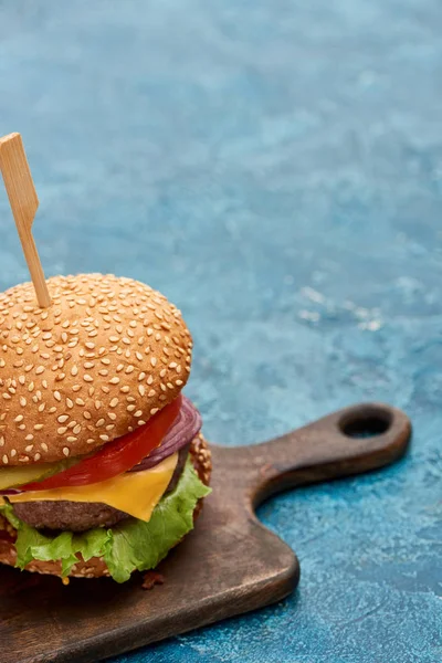 Köstlicher Cheeseburger auf Holzbrett auf blau strukturierter Oberfläche — Stockfoto