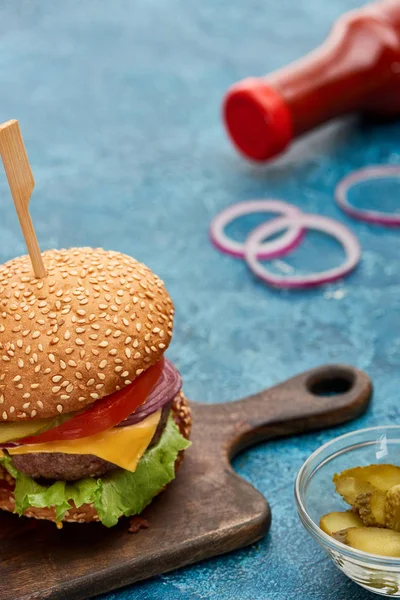 Selective focus of delicious cheeseburger on wooden board and pickles on blue textured surface — Stock Photo