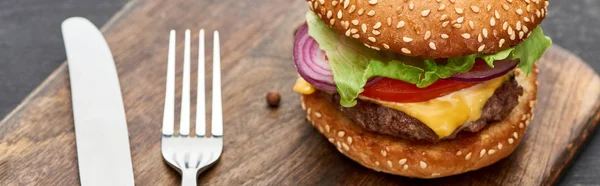Close up view of delicious fresh cheeseburger on wooden board with cutlery, panoramic shot — Stock Photo