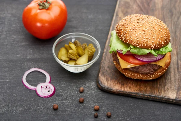 Deliciosa hamburguesa con queso fresco sobre tabla de madera cerca de tomate, encurtidos y aros de cebolla - foto de stock