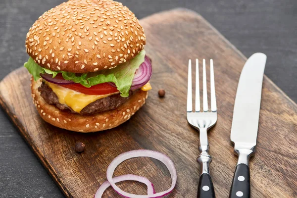 Delizioso cheeseburger fresco su tavola di legno con posate e anelli di cipolla — Foto stock