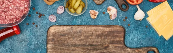Вид сверху на свежие ингредиенты бургера рядом с пустой разделочной доской на голубой текстурированной поверхности, панорамный снимок — стоковое фото