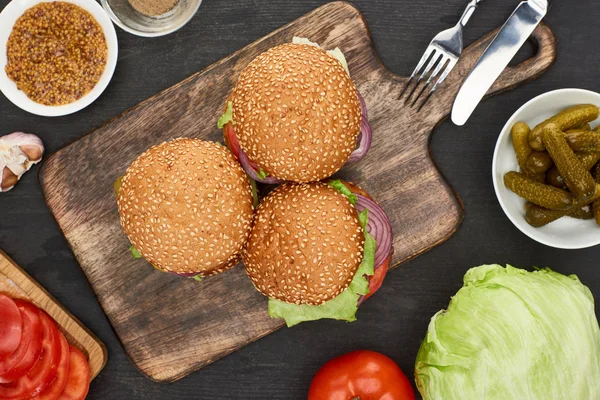 Vista superior de deliciosas hamburguesas frescas cerca de ingredientes y cubiertos en la tabla de cortar de madera - foto de stock