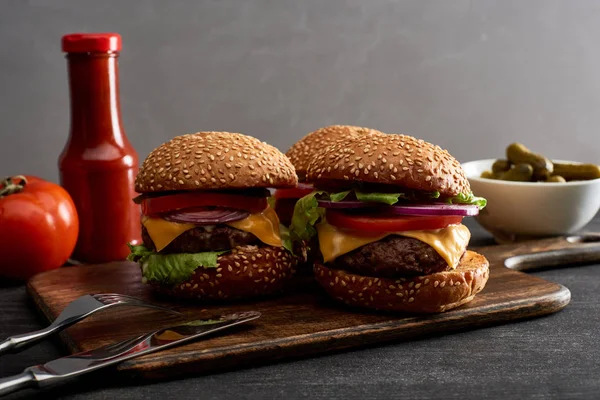 Delicious fresh meat cheeseburgers on wooden board near cutlery, ingredients and ketchup — Stock Photo