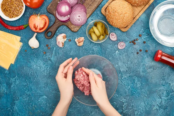 Vista superior da mulher segurando bawl com carne picada crua perto de ingredientes de hambúrguer fresco na superfície texturizada azul — Fotografia de Stock