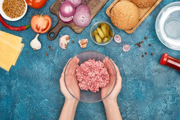 Vista superior da mulher segurando bawl com carne picada crua perto de ingredientes de hambúrguer fresco na superfície texturizada azul — Fotografia de Stock