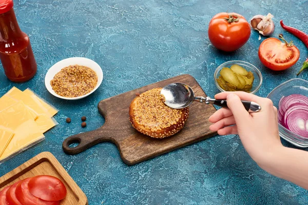 Cropped view of woman cooking delicious fresh burger with mustard on blue textured surface — Stock Photo