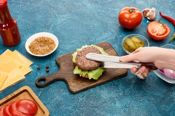 Vista recortada de la mujer cocinar deliciosa hamburguesa fresca en la superficie de textura azul - foto de stock