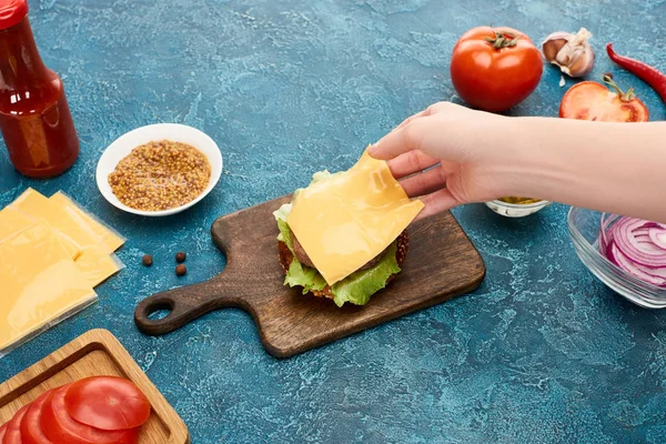Vue recadrée de femme cuisinant délicieux cheeseburger frais sur surface texturée bleue — Photo de stock
