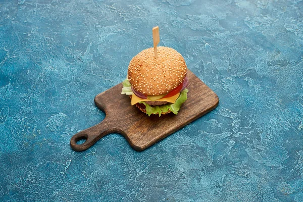 Delicioso cheeseburger na placa de madeira na superfície texturizada azul — Fotografia de Stock