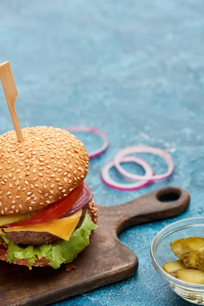 Selective focus of delicious cheeseburger on wooden board near pickles on blue textured surface — Stock Photo