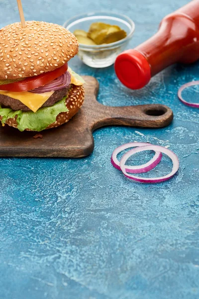 Selective focus of delicious cheeseburger on wooden board near pickles and ketchup on blue textured surface — Stock Photo