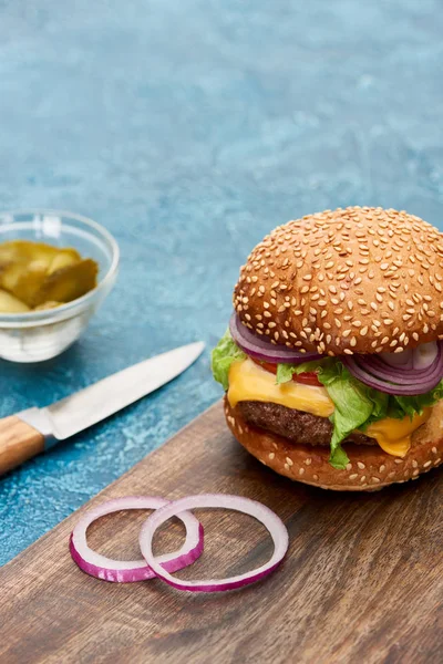Selective focus of delicious cheeseburger on wooden board near pickles and knife on blue textured surface — Stock Photo
