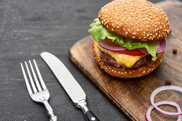 Delicious fresh cheeseburger on wooden board near cutlery — Stock Photo