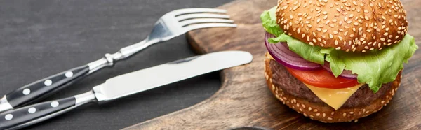 Delicious fresh cheeseburger on wooden board near cutlery, panoramic shot — Stock Photo