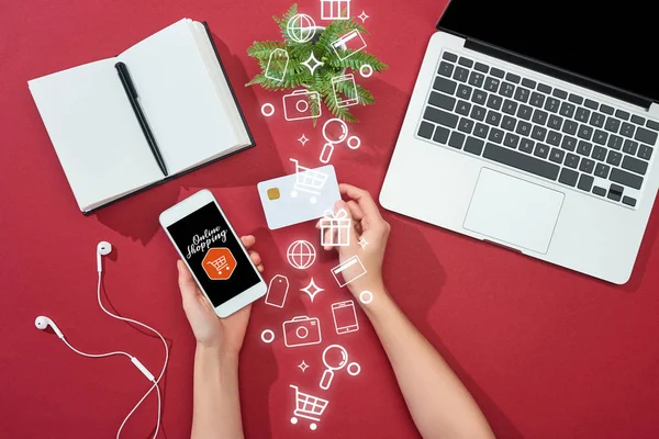 Обрезанный вид женщины, держащей кредитную карту и смартфон с иллюстрацией покупок онлайн рядом с ноутбуком, наушниками, ручкой, ноутбуком и растением на красном фоне — стоковое фото