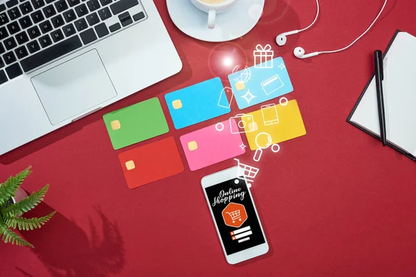 Вид зверху на різнокольорові порожні кредитні картки на червоному тлі з ілюстрацією інтернет-магазинів на смартфоні біля ноутбука, навушники та кава — стокове фото