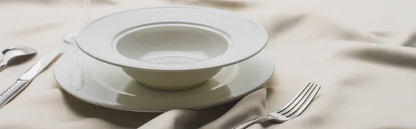 Tiro panorâmico de pratos e talheres em toalha de mesa branca ondulada — Fotografia de Stock