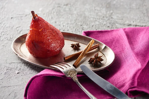 Deliziosa pera in vino con cannella e anice su piatto d'argento su superficie di cemento grigio con tovagliolo rosa, coltello e forchetta — Foto stock