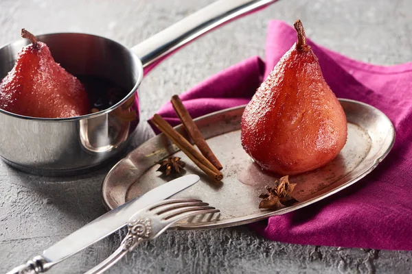 Delicioso pera en vino con canela y anís en plato de plata y en olla de estofado en superficie de hormigón gris con servilleta rosa, cuchillo y tenedor - foto de stock