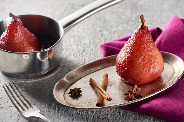 Deliziosa pera in vino con cannella e anice su placca d'argento e in pentola su superficie di cemento grigio con tovagliolo rosa e forchetta — Foto stock