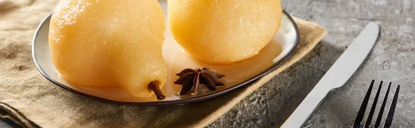 Delicious pear in wine with anise and cinnamon served on plate on napkin with knife and fork on grey concrete surface, panoramic shot — Stock Photo