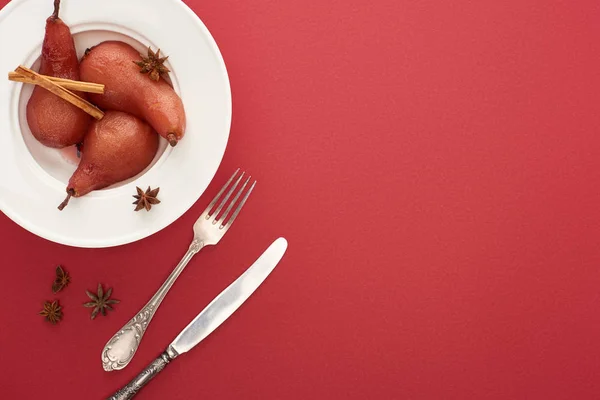 Top view of delicious pear in wine with cinnamon and anise on plate on red background with cutlery — Stock Photo