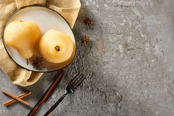 Vista dall'alto di deliziose pere in vino con anice e cannella servite su piatto su tovagliolo con coltello e forchetta su superficie in cemento grigio — Foto stock