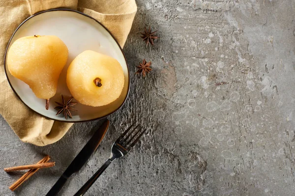 Top view of delicious pear in wine with anise and cinnamon served on plate on napkin with knife and fork on grey concrete surface — Stock Photo