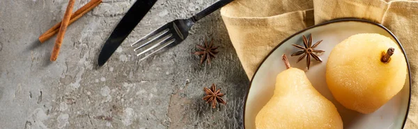 Vista dall'alto di deliziosa pera in vino con anice e cannella servita su piatto su tovagliolo con coltello e forchetta su superficie in cemento grigio, colpo panoramico — Foto stock