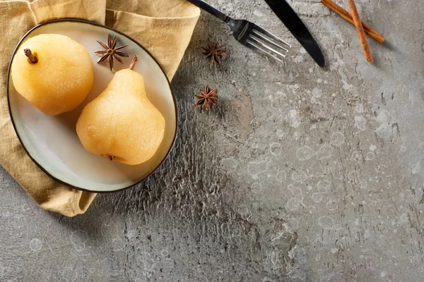 Vue de dessus de délicieuse poire dans le vin à l'anis et à la cannelle servie dans une assiette sur une serviette avec couteau et fourchette sur une surface en béton gris — Photo de stock