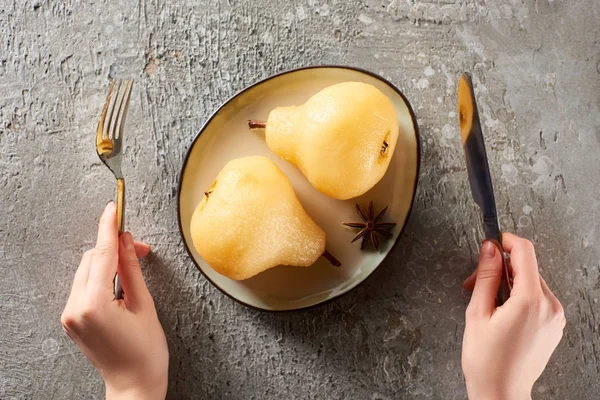 Cropped view of woman holding fork and knife near delicious pear in wine on grey concrete surface — Stock Photo