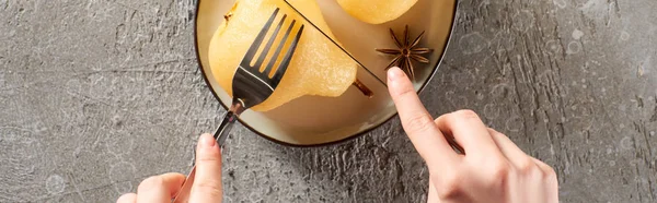 Vista recortada de la mujer comiendo deliciosa pera en vino con tenedor y cuchillo en la superficie de hormigón gris, plano panorámico - foto de stock