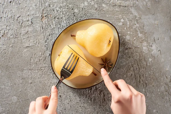 Cropped view of woman eating delicious pear in wine with fork and knife on grey concrete surface — Stock Photo