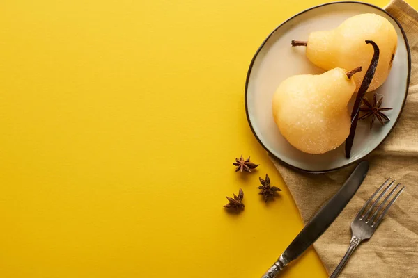 Vista superior de la deliciosa pera en vino con anís en el plato cerca de cubiertos y servilleta sobre fondo amarillo - foto de stock