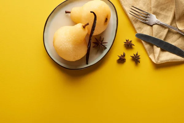 Vista superior de la deliciosa pera en vino con anís en el plato cerca de cubiertos y servilleta sobre fondo amarillo - foto de stock