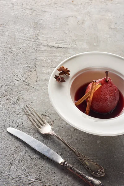 Deliziosa pera in vino rosso con cannella e anice su piatto su superficie di cemento grigio con posate — Foto stock