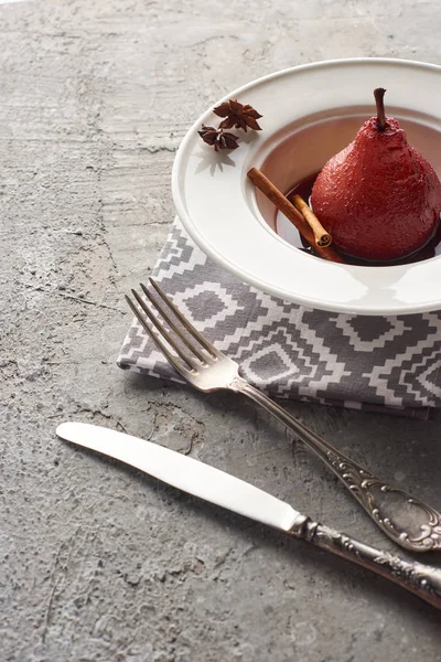 Deliciosa pera en vino tinto con canela y anís en plato sobre superficie de hormigón gris con cubiertos en servilleta - foto de stock