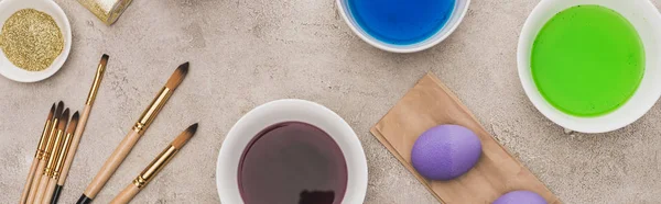 Top view of chicken eggs, glitter and watercolor paints with paintbrushes on grey concrete surface, panoramic shot — Stock Photo