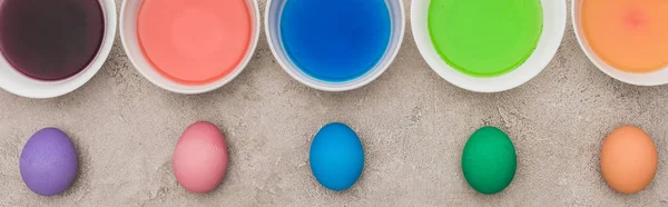 Top view of painted chicken eggs near watercolor paints in bowls on grey concrete surface, panoramic shot — Stock Photo