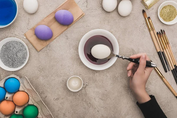 Vista recortada de la mujer para colorear huevos de pollo en pinturas de acuarela en la superficie de hormigón gris - foto de stock