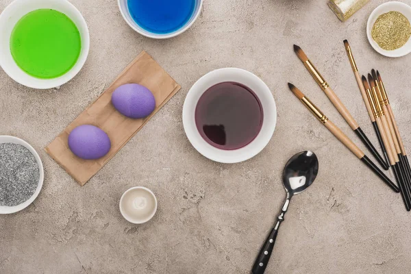 Top view of chicken eggs, glitter and watercolor paints with paintbrushes on grey concrete surface — Stock Photo