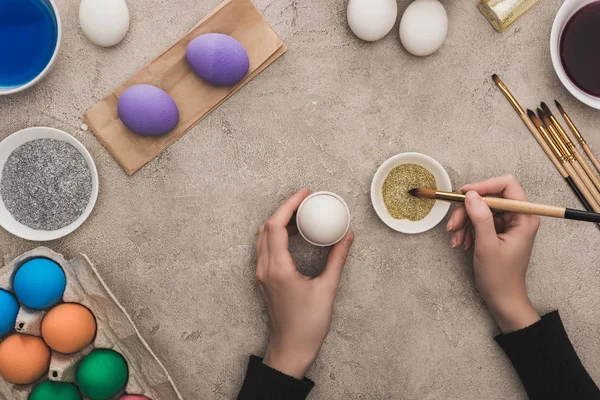 Vista ritagliata della donna che decora le uova di pollo con brillantini dorati sulla superficie di cemento grigio — Foto stock
